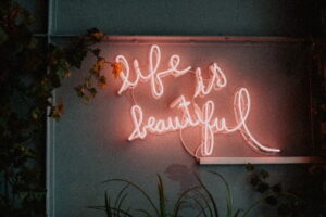 Neonbord met de woorden Life is Beautiful tegen een muur met groene planten — symbool voor dankbaarheid, positiviteit en gelukkig zijn in het moment.