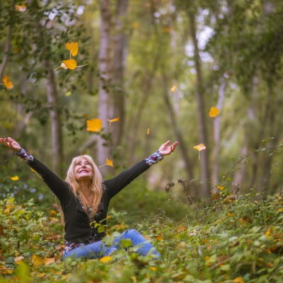 Blonde vrouw zit vrolijk tussen de herfstbladeren in een bos en gooit de bladeren omhoog — symbool voor vrijheid, loslaten en gelukkig zijn in het moment.